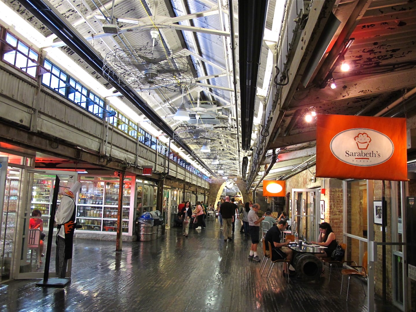 A bustling indoor food hall with shared tables, warm lighting, and visitors drying off umbrellas near the entrance