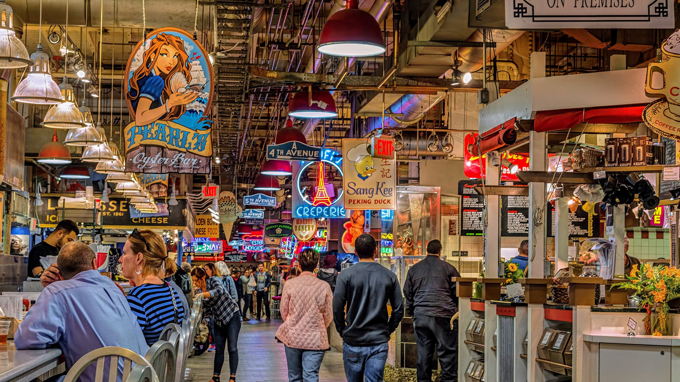 A busy indoor market aisle with food counters on both sides and warm hanging lights over a crowd of shoppers