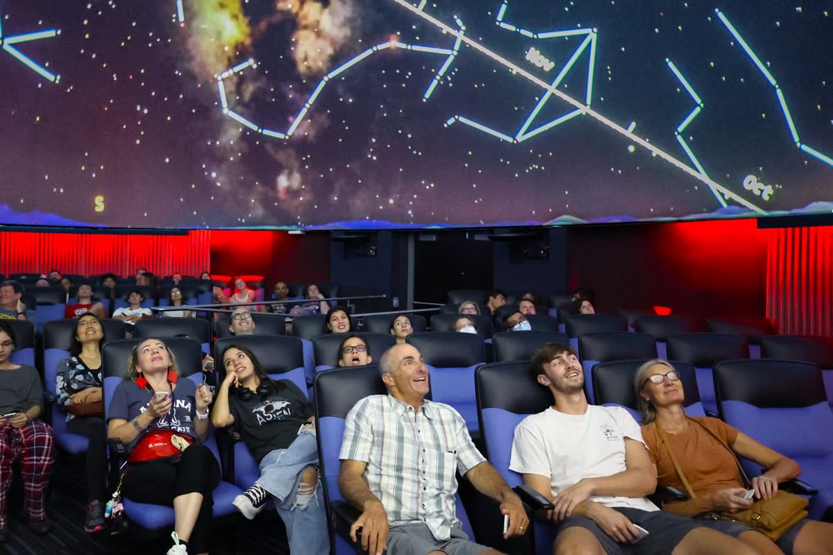 A family sitting together in a planetarium theater, heads tilted up toward a projected night sky as the dome glows softly