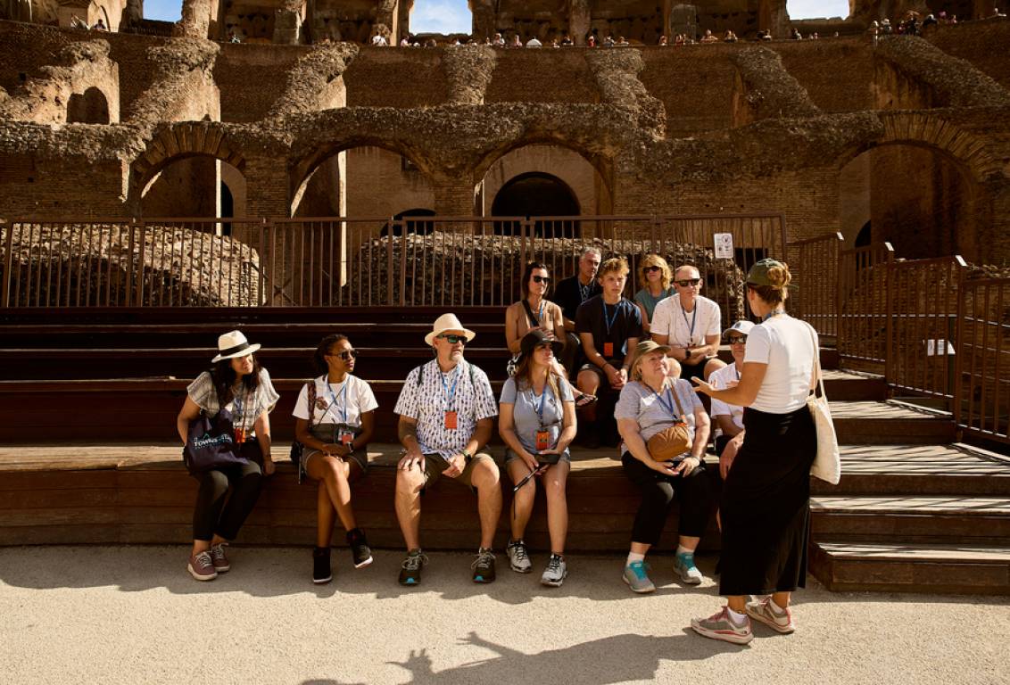 A guide speaking to a small group inside the Roman Colosseum, with the stone arches and arena floor visible behind them in bright daylight