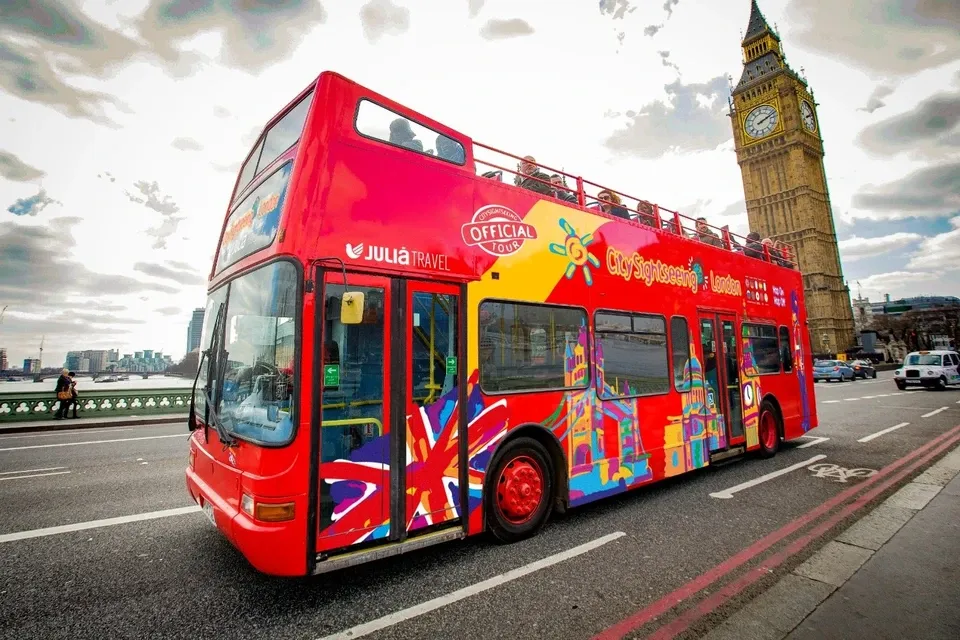 A hop-on hop-off sightseeing bus passing a busy city street with tourists seated on the open top deck and a famous landmark visible in the distance