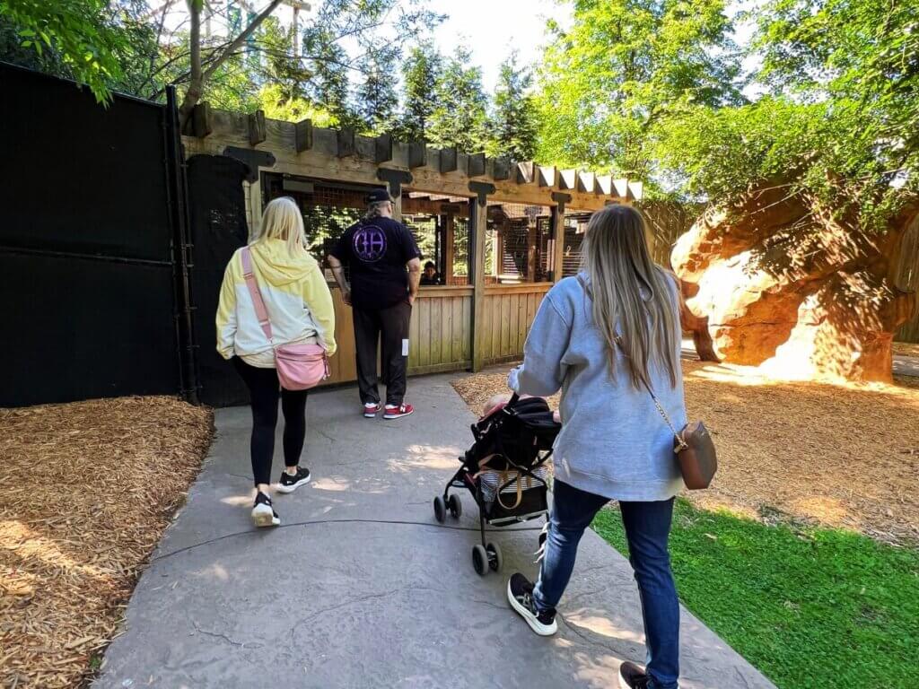 A parent pushes a stroller along a wide zoo pathway lined with trees near exhibit signs.