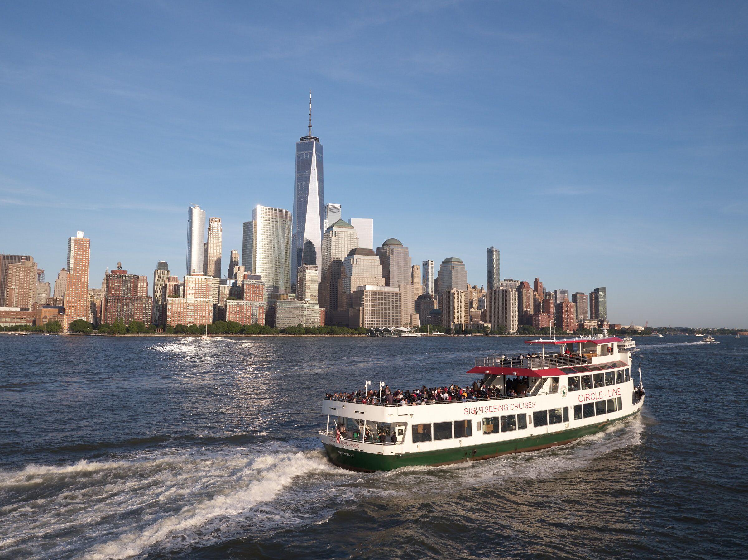 A sightseeing boat in New York Harbor with the Manhattan skyline behind it as passengers lean on the railings