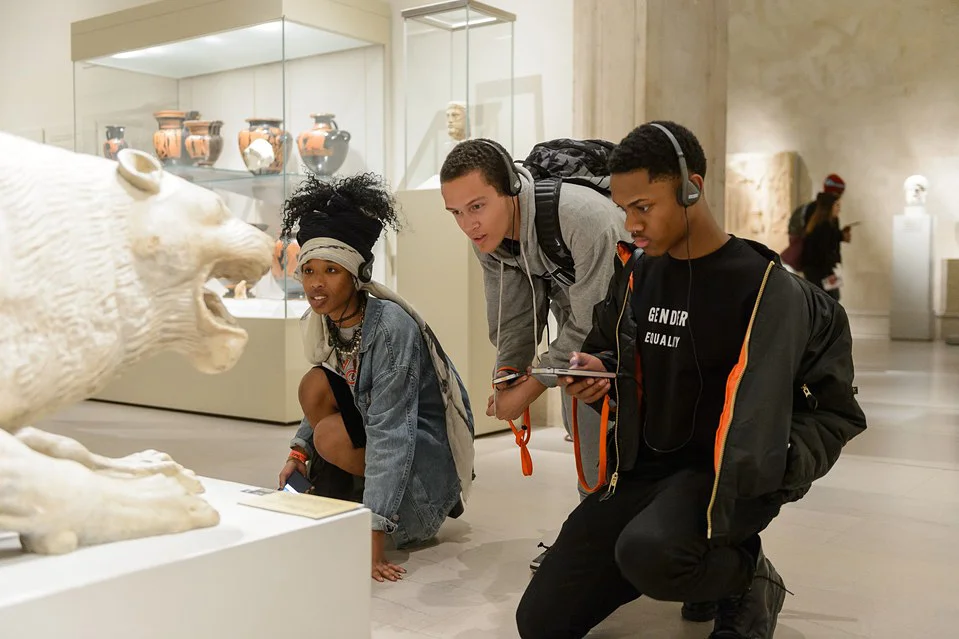 A visitor holding a smartphone with an audio guide screen open while standing in a museum gallery with sculptures and visitors in the background
