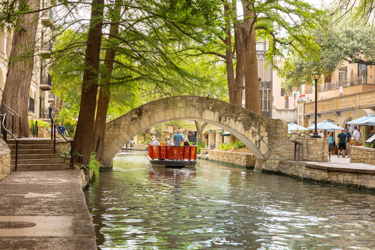 A wide, gently curving riverside walkway with smooth paving, railings, and shaded seating areas beside the water