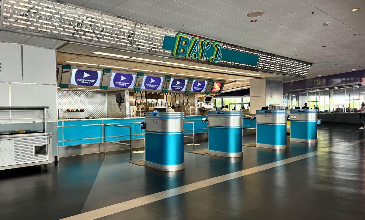 Guests ordering at a theme park quick-service counter with menu boards overhead and a visible queue area