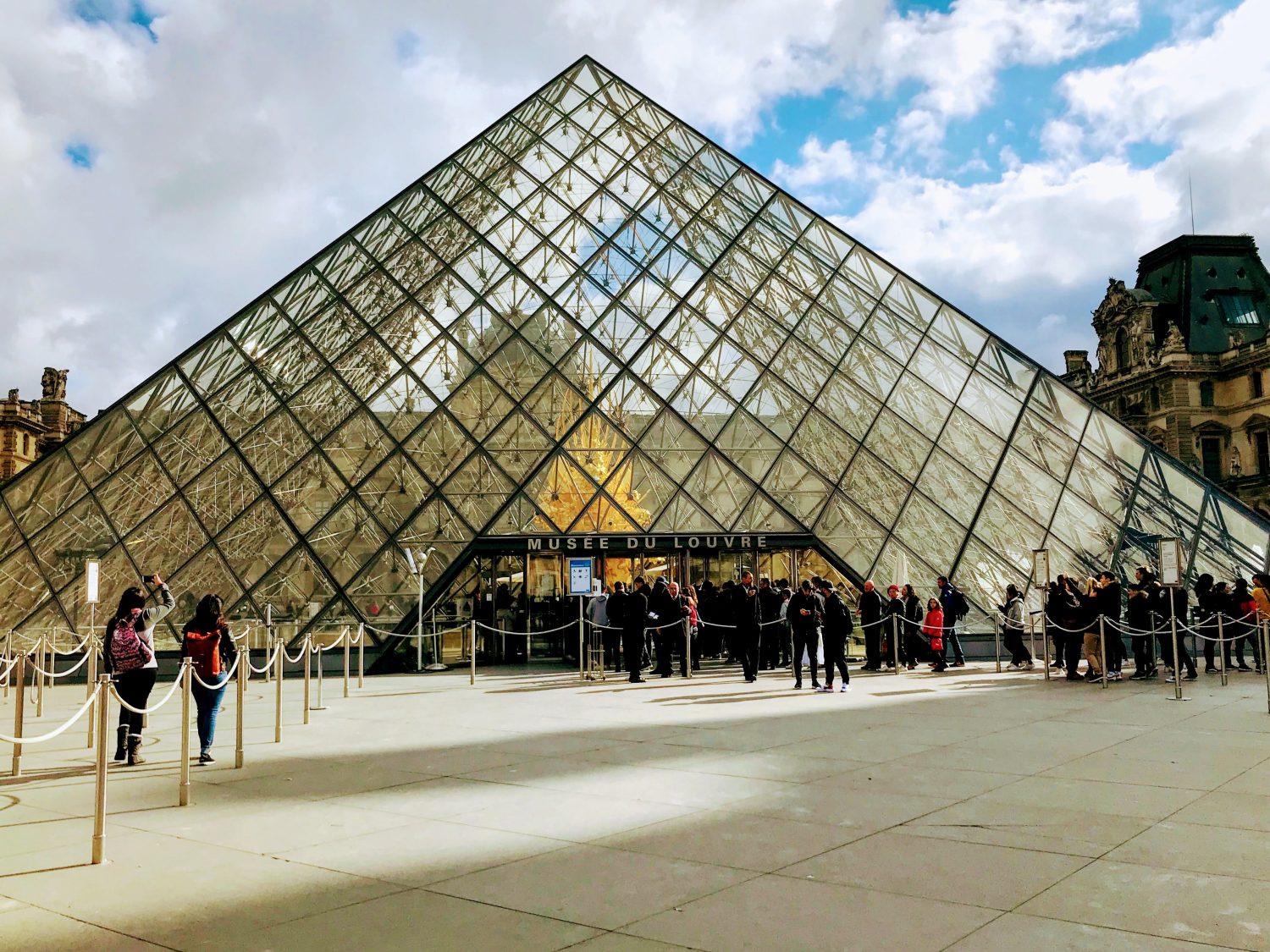 Travelers waiting in a line outside a well-known museum entrance on a sunny day, with timed-entry signs and people checking tickets on their phones